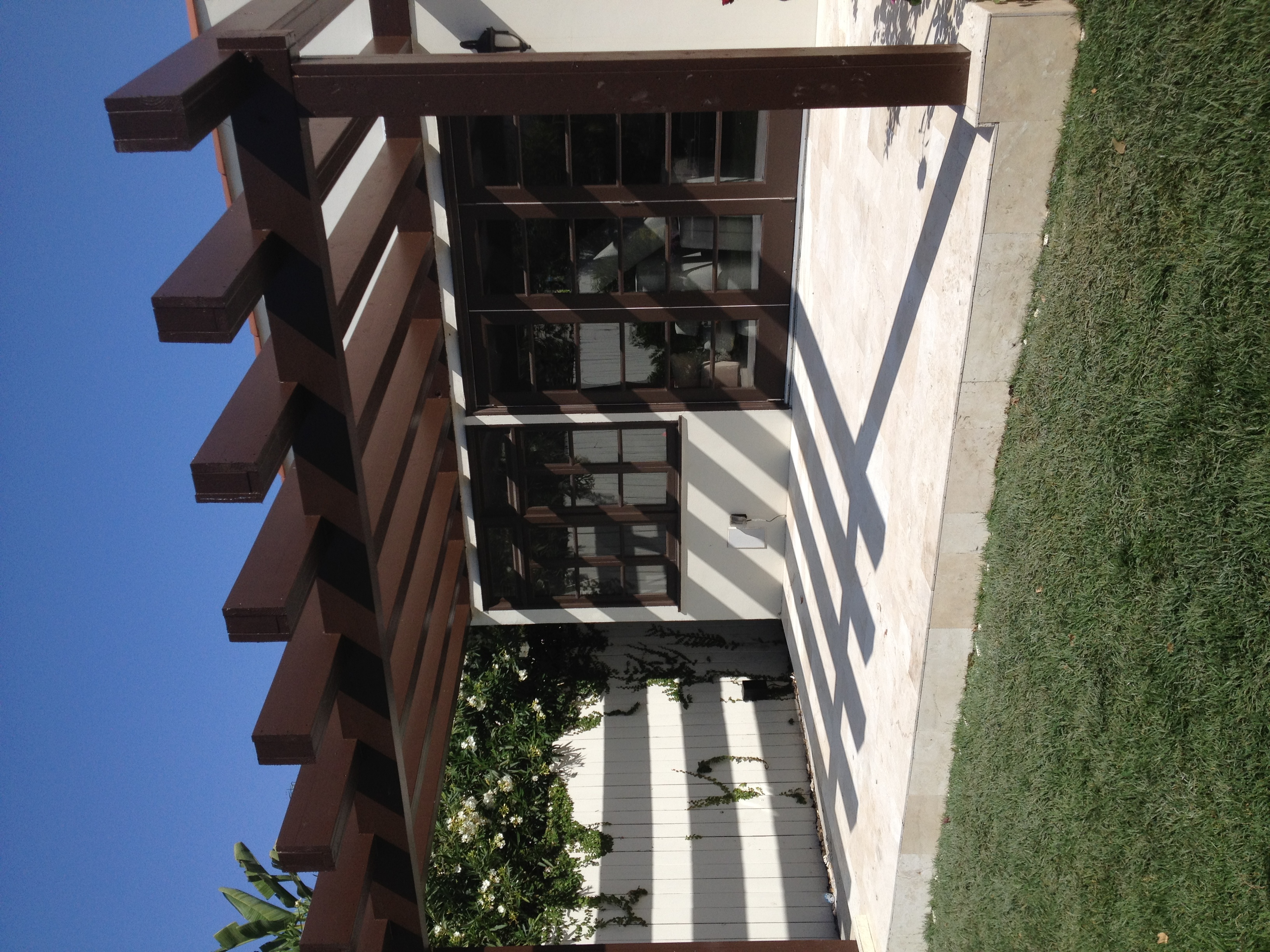 Custom wood pergola casting dramatic shadows over travertine stone patio, blue sky above, luxury residential exterior construction by PBG.